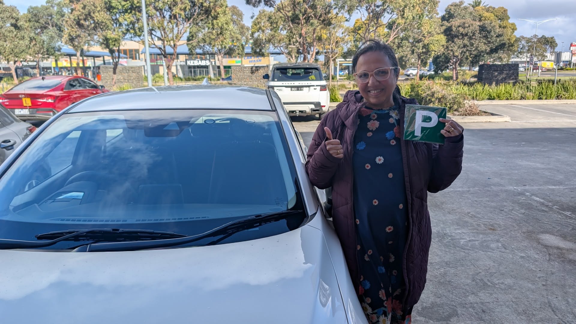 Learner holding a green P plate and giving a thumbs up beside a white car