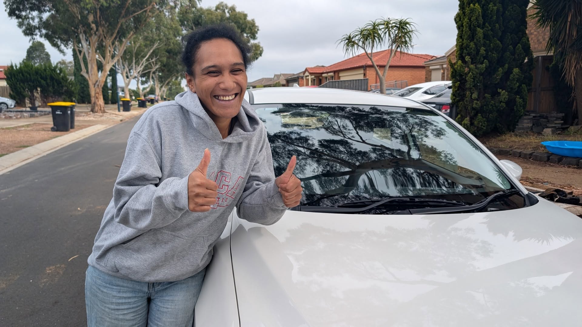 Learner giving two thumbs up beside a white car on a suburban street
